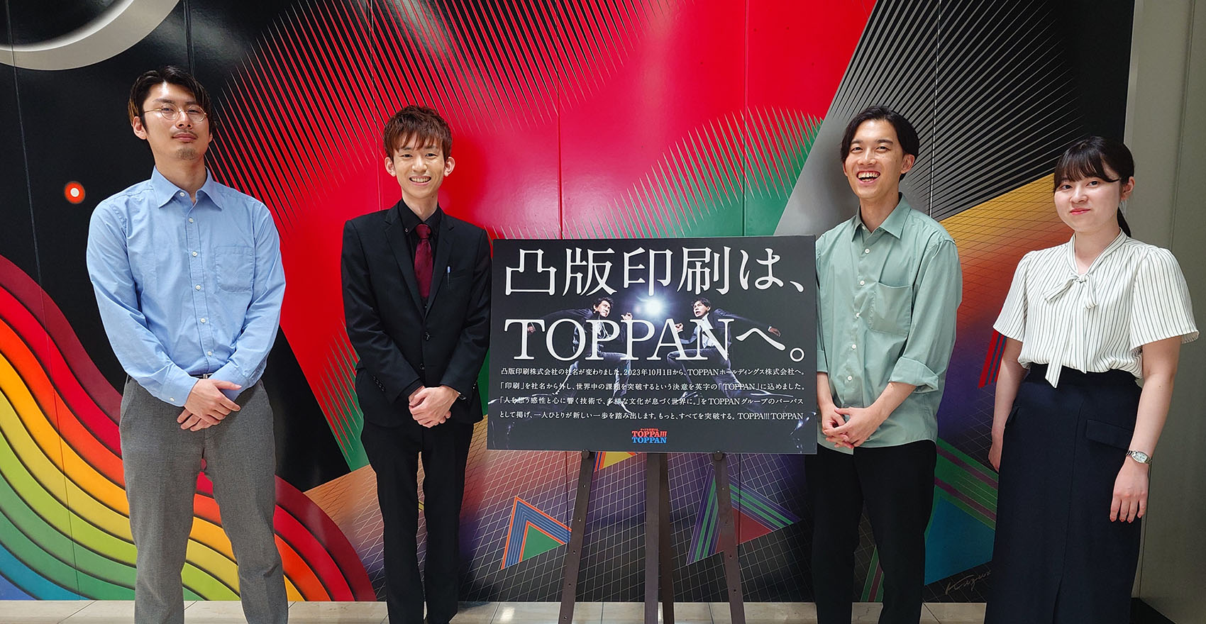 男性社員3名と女性社員1名が「凸版印刷は、TOPPANへ」の看板とともに横一列に並び、笑顔をむける様子