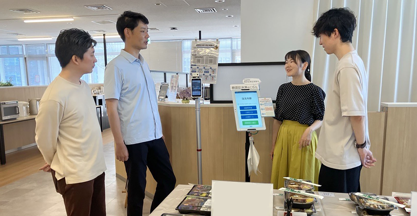 弁当が並んだテーブルの前で、男性社員3名に向かってシステムについて説明する女性社員1名